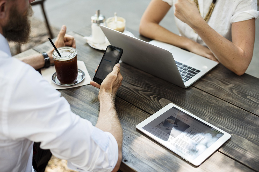 a man and a woman sitting at a wooden table outside, the man has a glass of a brown liquid with a straw and is looking at his cell phone, the woman is sitting in front of an opened laptop computer
